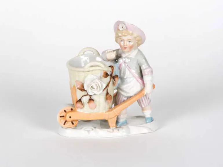 Sculpture en porcelaine IB05062 d’un jeune enfant avec une charrette et un panier, peinte et émaillée, sur fond blanc.