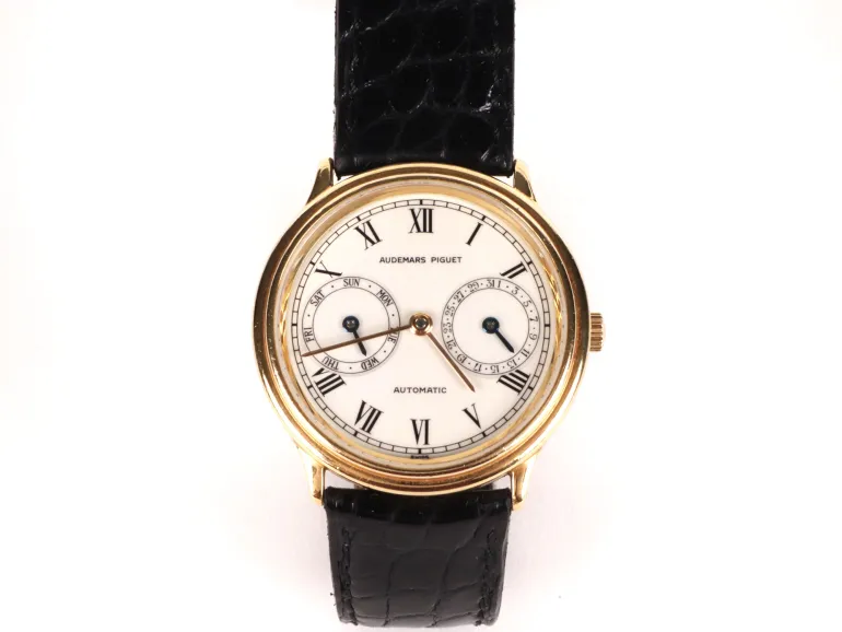 Montre Audemars Piguet Classic Day Date, SKU IB09473, boîtier en or, cadran blanc à chiffres romains, sous-cadrans jour et date, bracelet noir façon croco.