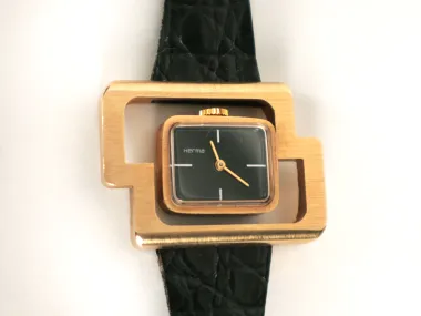 Herma watches IB09261 vintage 1970s wristwatch with gold-tone asymmetrical case, black rectangular dial, and black strap.