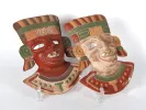 Two painted terracotta Aztec masks, IB04480, in red and cream with green and ochre details on display stands.
