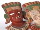 Two painted terracotta Aztec-style masks, one red and one cream, with green and ochre details, SKU IB04480.