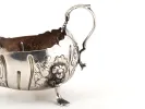 IB09116 18th-century Irish sterling silver sauceboat with one handle, ornate floral and figure decoration, and three feet; shown in side view.